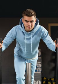 Felpa azzurra e pantaloni jogger coordinati realizzati in tessuto morbido. L'individuo si appoggia a una barra di metallo in un ambiente di palestra, con attrezzi da allenamento sullo sfondo.