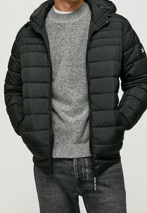 Mężczyzna w czarnej pikowanej kurtce zapinanej na zamek błyskawiczny z kapturem, szarym swetrze i ciemnoszarych dżinsach, z rękami w kieszeniach kurtki.