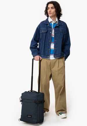 Dark blue fabric backpack with a rectangular design, adjustable straps, and a front zip pocket, carried alongside a wheeled suitcase.