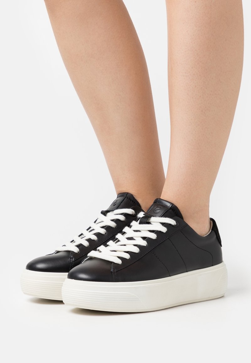 Baskets en cuir noir avec des lacets blancs, présentant une semelle en caoutchouc texturée et un dessus lisse. Design bas, bout arrondi, accents minimaux.