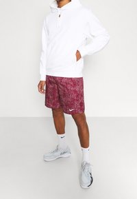 Sweat à capuche blanc avec poche avant et cordon de serrage, associé à un short maron chiné avec un logo Nike blanc. Baskets gris clair et chaussettes blanches.