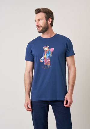 Man met baard die een marineblauw T-shirt draagt met een kleurrijke beer in skikleding die een snowboard vasthoudt, staand tegen een eenvoudige achtergrond.