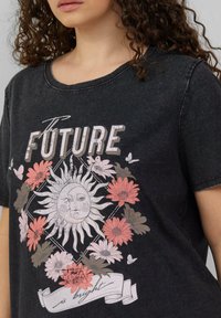 Zwarte katoenen T-shirt met korte mouwen, voorzien van een zonnenafbeelding en bloemen in roze en grijs met de tekst "De TOEKOMST is helder."