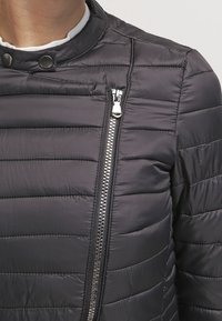 Chaqueta negra acolchada con cuello alto, costuras horizontales y un bolsillo con cremallera plateada en el lado izquierdo. Textura suave.