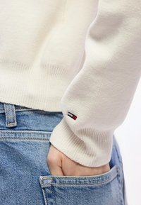 Άτομο που φοράει κρεμ πουλόβερ με μικρό κόκκινο, λευκό και μαύρο λογότυπο στο μανίκι, το χέρι ακουμπάει στην μπροστινή τσέπη μπλε τζιν παντελονιού.