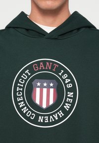 Zelena dukserica s kapuljačom s okruglim grafičkim dizajnom koji sadrži natpis "GANT 1949 NEW HAVEN CONNECTICUT" i štitni dizajn s crvenim i bijelim prugama i zvijezdama.