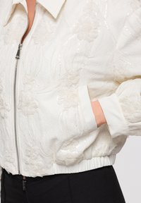Veste en dentelle florale blanche avec fermeture éclair, ourlet et poignets élastiques, portée avec une main dans la poche latérale sur un pantalon noir.