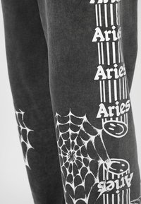 Szare bawełniane spodnie dresowe z białymi nadrukami pajęczyny i napisem "Aries" po bokach. Materiał wydaje się miękki o gładkiej fakturze.