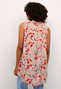 Mouwloze blouse in beige met een bloemmotief in rood en roze. Heeft een gerimpelde kraag en een gebogen zoom. Relaxte pasvorm.