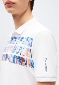 Νέος άνθρωπος που φοράει λευκό πόλο Calvin Klein με πολύχρωμες αφηρημένες ρίγες στο στήθος και το όνομα της μάρκας στο μανίκι.