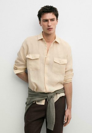 Man die een beige overhemd met lange mouwen en twee borstzakken draagt en een olijfkleurige trui om zijn middel heeft gebonden, staand tegen een witte achtergrond.