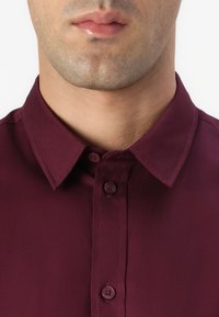 Bordowa koszula z guzikiem i klasycznym kołnierzykiem, z dwoma guzikami na plisie, wykonana z gładkiego materiału o subtelnym połysku.