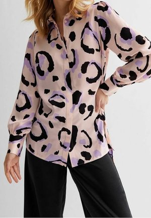 Femme portant un chemisier rose clair avec des taches abstraites de léopard noires et lavande et un pantalon noir, posant avec une main sur la hanche.
