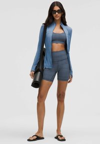 Chaqueta deportiva azul claro, top de cuadros y pantalones cortos, esterilla de yoga negra en la mano, combinada con chanclas negras, diseño elegante, material flexible.