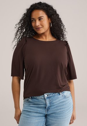 Femme aux cheveux bouclés portant des boucles d'oreilles créoles argentées, un haut marron à manches froncées et un jean bleu clair sur un fond uni.