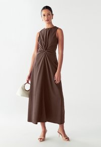 Vestido marrón sin mangas con un detalle frontal retorcido, que presenta una silueta de largo hasta el suelo, confeccionado en una tela suave. Accesorio: bolso blanco.