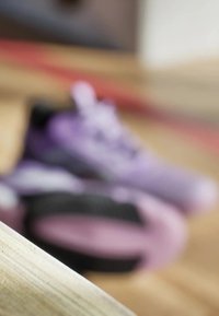Purple athletic shoes with a soft texture and contrasting black accents, partially blurred in the background against a wooden floor.