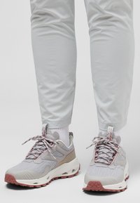 Baskets de sport grises avec un tissu texturé, des accents rouges et une semelle épaisse et à motifs, assorties à un pantalon gris clair et des chaussettes blanches.