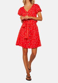 Robe fleurie rouge avec un col en V, des manches courtes, des boutons sur le devant et une ceinture en tissu à la taille. Matière légère et texturée.