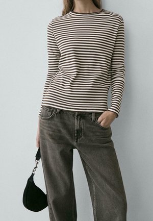 Personne portant une chemise à manches longues rayée marron et blanche et un jean gris foncé, tenant une petite pochette noire à côté d'elle.