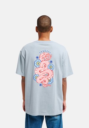 Mann trägt hellblaues T-Shirt mit großem pinkem Schlangen- und blauem Blumengrafik auf dem Rücken, kombiniert mit blauen Jeans, steht mit dem Rücken zur Kamera.
