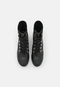 Musta nahkast korkeat kengät, joissa on kultaiset silmukat ja nauhat. Kenkien kärki on kiinteä mustasta kumista ja niissä on teksturoitu kangas yksityiskohtana.