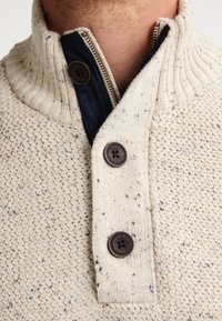 Pullover a maglia color crema con colletto alto, presenta accenti di bottoni marroni e uno strato di denim visibile all'area del colletto e della zip.