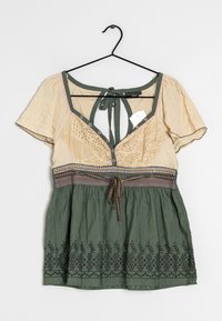 Blouse beige et verte avec motif floral brodé, manches courtes, lien à nouer dans le dos et fermeture par bouton sur un tissu texturé sur le haut.