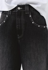 Jean noir taille haute avec des clous argentés le long des poches avant et des surpiqûres blanches contrastées, porté avec un haut blanc rentré.