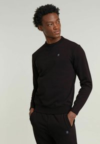 Felpa nera realizzata in tessuto morbido con collo rotondo, polsini a costine e un piccolo logo sul petto. Indossata con pantaloni abbinati.