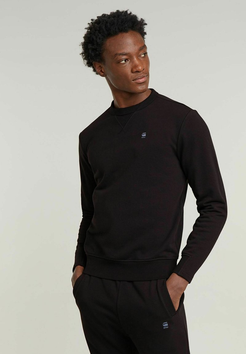 Felpa nera realizzata in tessuto morbido con collo rotondo, polsini a costine e un piccolo logo sul petto. Indossata con pantaloni abbinati.