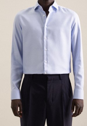 Uomo che indossa una camicia azzurra chiaro a maniche lunghe con trama, infilata in pantaloni plissettati a vita alta di colore blu navy scuro.