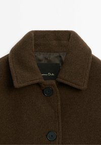 Brauner Wollmantel mit einem auffälligen Kragen, schwarzen Knöpfen und weicher Textur. Etikett innen sichtbar, keine zusätzlichen Muster oder Akzente.