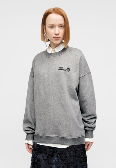 Γκρι φούτερ με χαλαρή εφαρμογή και ριμπ γιακά, με κεντημένο το κείμενο "BAUM UND PFERDGARTEN" σε μαύρο. Φοριέται πάνω από ένα λευκό πουκάμισο με γιακά.