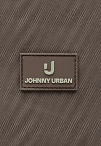 Hnedá texturovaná látka s prišitým obdĺžnikovým štítkom s nápisom "JOHNNY URBAN" pod štýlizovaným logom "J" rovnakej farby.