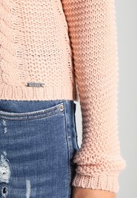 Maglione lavorato a maglia di un rosa chiaro con un design a trecce testurizzato, orlo corto e polsini a costine. Presenta un'etichetta con il logo "GUESS" in argento. Abbinarlo a jeans strappati.