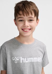 Tricou din bumbac gri cu guler rotund și un logo imprimat alb. Design simplu, cu mâneci scurte și croială normală.