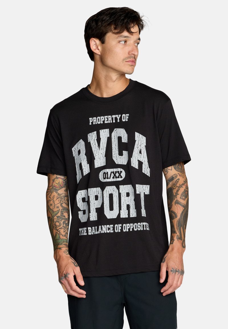 Giovane uomo con tatuaggi che indossa una maglietta nera con testo bianco che recita "PROPRIETÀ DI RYCA SPORT L'EQUILIBRIO DEGLI OPPOSTI."