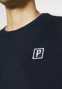 Tmavě modré bavlněné tričko s kulatým výstřihem a bílým vyšitým logem v čtverci na levé části hrudi. Hladká textura.