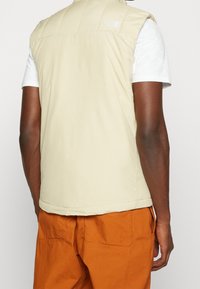 Beige gepolsterte Weste mit hohem Kragen und gesticktem Logo auf dem Rücken, getragen über einem weißen Shirt, kombiniert mit orangefarbenen Hosen.