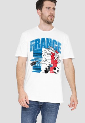 Man wearing white t-shirt with blue "FRANCE" text and Bugs Bunny kicking a soccer ball in red, white, and blue design, paired with blue jeans.