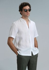 Chemise blanche en lin à manches courtes avec un devant à boutons et un col, portée avec un pantalon vert olive. Texture lisse, coupe décontractée.