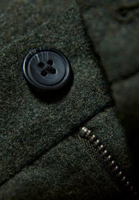 Ciemnozielona wełniana tkanina z czarnym guzikiem z czterema otworami i srebrnym zamkiem błyskawicznym, podkreślająca fakturę i funkcjonalne detale.