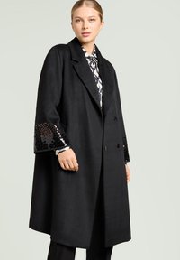 Svart yllekappa med dubbelknäppt design. Har paljettdekorationer på ärmsluten. Mjuk textur, breda slag och en avslappnad silhuett.