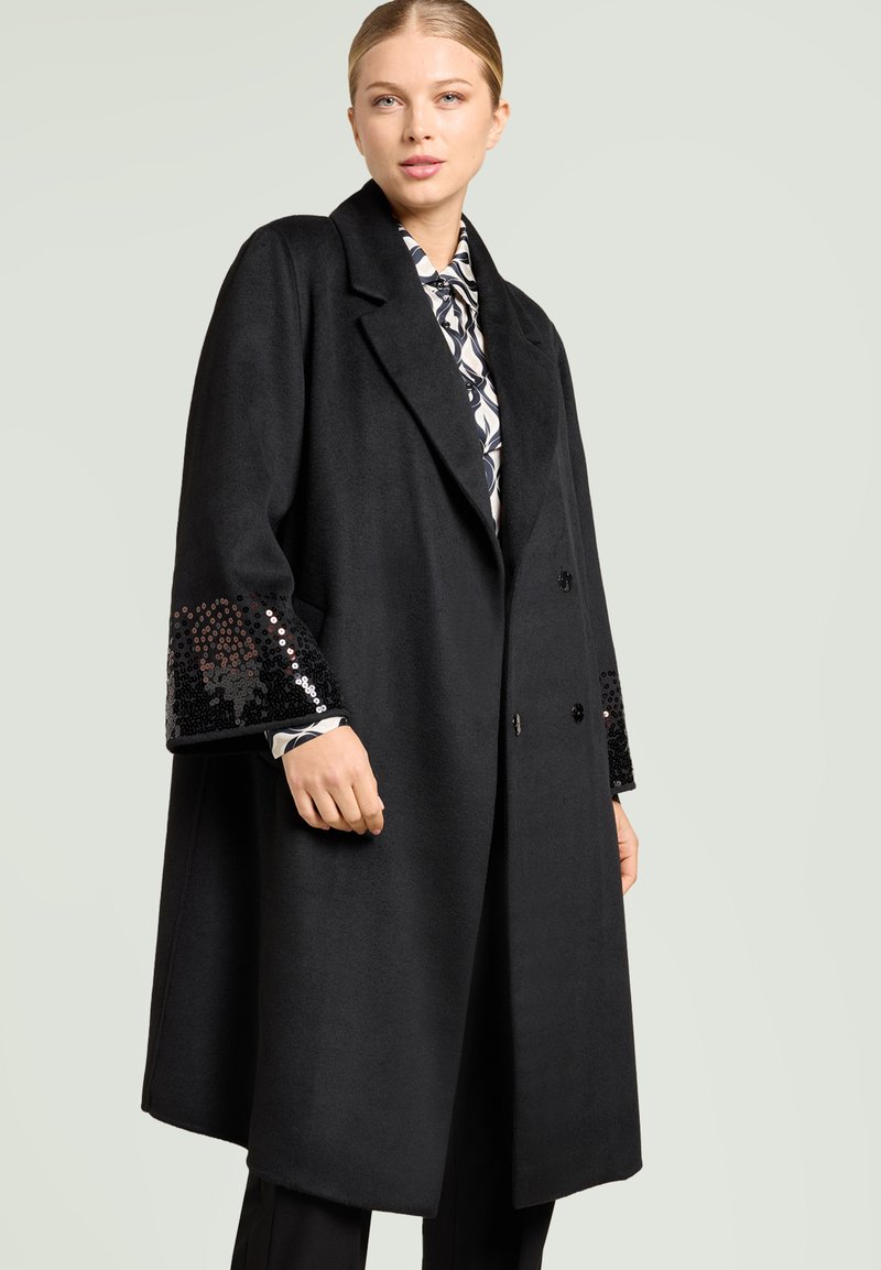 Svart yllekappa med dubbelknäppt design. Har paljettdekorationer på ärmsluten. Mjuk textur, breda slag och en avslappnad silhuett.