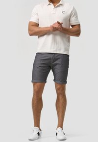 Polo skjorte i lys beige, korte ærmer, med et lille logo. Grå denim shorts, opbrudte kanter, parret med hvide sneakers med sorte accenter.