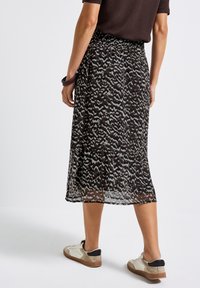 Personne portant une jupe midi à motifs noirs et gris, un haut marron, des baskets blanches et un bracelet sombre, se tenant sur un sol blanc.