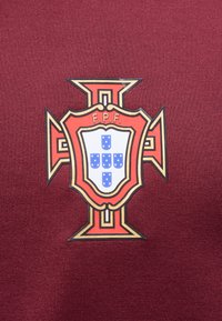 Emblème de croix rouge avec des contours orange, noir et or entourant un bouclier blanc avec cinq petits boucliers bleus et les lettres "F.P.F." sur un tissu bordeaux.