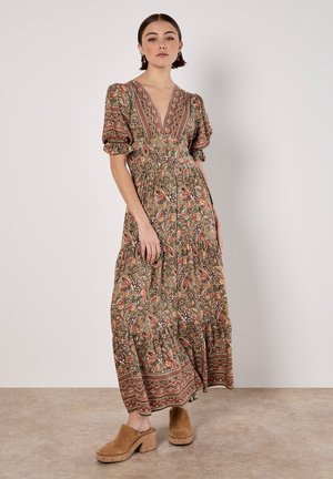 Langes, gemustertes Kleid in erdigen Tönen mit Puffärmeln, tiefem V-Ausschnitt und gestuftem Rock. Getragen mit braunen, klobigen Clogs mit Absatz.