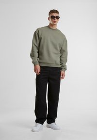 Grøn crewneck sweatshirt med ribbede manchetter, parret med sorte vidde bukser og hvide sneakers; model iført mørke solbriller.